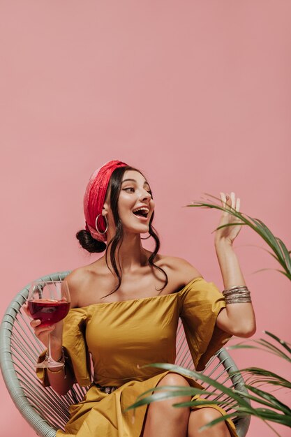 Free Photo emotional trendy woman in bright headband, silver earrings and yellow cool dress greeting and holding glass with cocktail