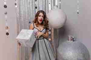 Free photo emotional white woman in long party dress posing with amazement, holding big present box. winsome female model in sparkle attire standing near huge christmas toys with gift.