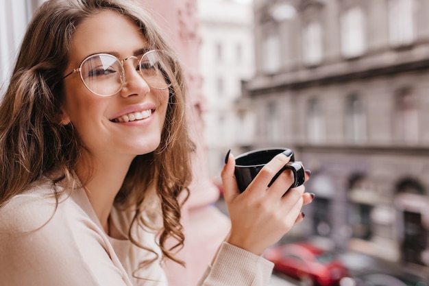 Emotional white woman wears glasses posing on blur background with cup of hot beverage