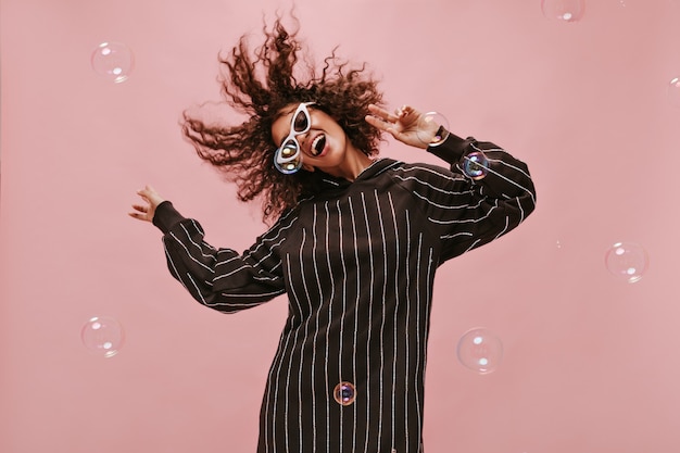 Free photo emotional woman in modern black dress and sunglasses playing her wavy hair, smiling and dancing on isolated pink wall..