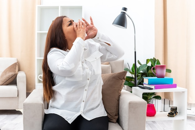 Emotional woman in white shirt and black pants sitting on the chair shouting with hands near mouth in light living room