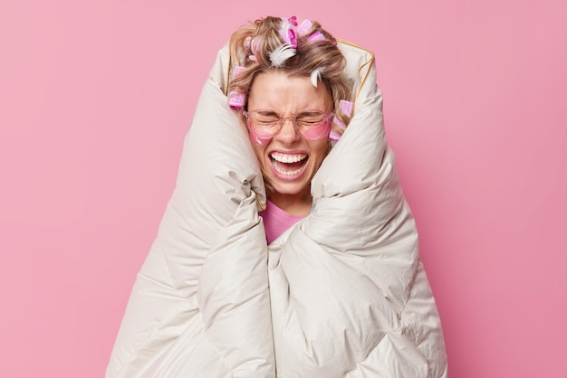 Emotional woman yells loudly wrapped in soft blanket wears transparent eyeglasses hydrogel patches makes hairstyle with rollers poses in bedroom against pink background. Morning daily routines