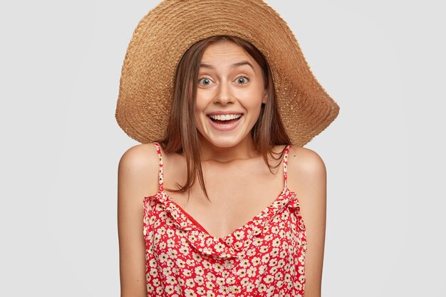 Emotional young lady has joyful expression, wears summer straw hat and fashionable dress