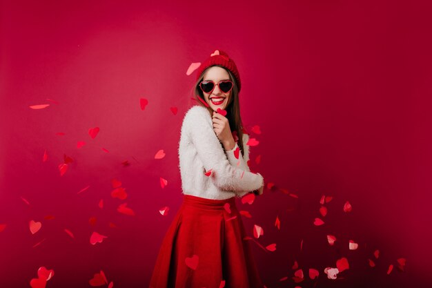 Emotional young woman in red hat and sunglasses standing on claret space at party