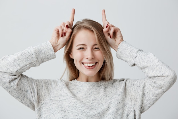 Free photo emotional young woman in sweater smiling broadly, mocking, making faces, holding index fingers over her head