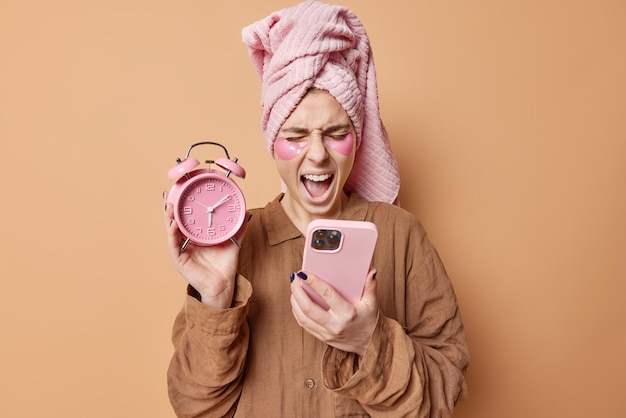 Emotional young woman yells loudly looks at smartphone checks receives message being in hurry as awakes late holds alarm clock hates early awakening dressed in nightwear wrapped towel on head