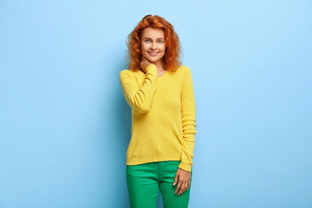 Emotions concept. Charming lovely redhaired woman touches neck and smiles pleasantly at camera