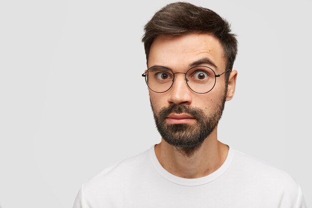 Emotive bearded young guy with thick stubble, trendy hairdo, looks with surprise