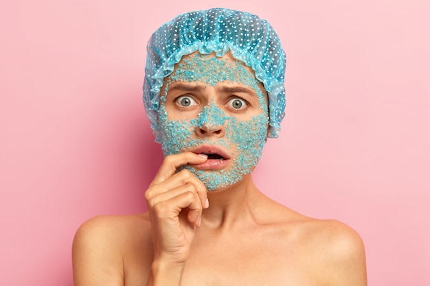 Free photo emotive dissatisfied woman looks with unhappy expression, keeps fore finger on lips, hears bad news from beautician, applies nourishing blue sea salt mask