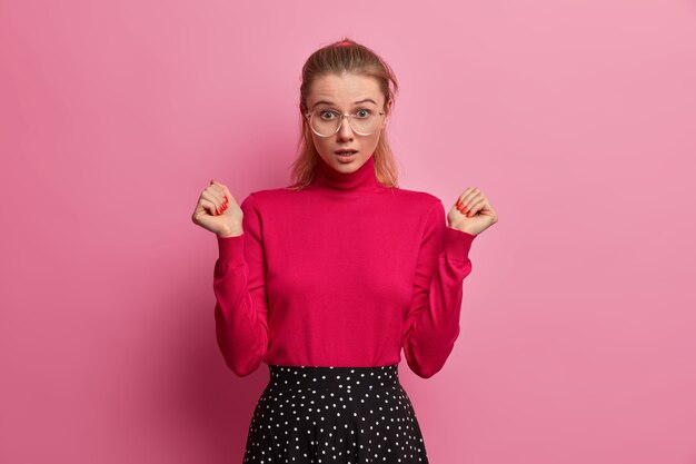 Emotive young adult female holds breath, hears something astonishing, raises hands, cannot believe her eyes, wears turtleneck and skirt, sees amazed thing