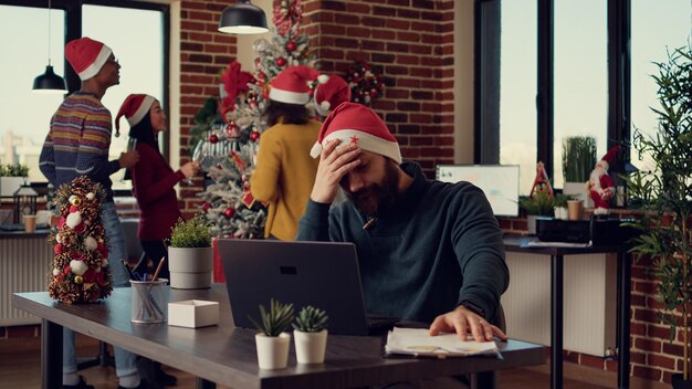 Employee being frustrated by christmas holiday festivity in startup office, trying to work on laptop during xmas eve time. Stressed man being disturbed and interrupted by noisy people.