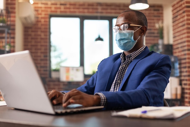 Employee with face mask using laptop to work on project planning and business strategy. Person working on computer to do paperwork and work on startup company during covid 19 pandemic.