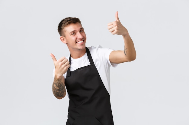 Employees, grocery stores and coffee shop concept. Handsome friendly barista in black apron, showing thumbs-up, inviting for cappuccino, smiling delighted, guarantee quality, white background