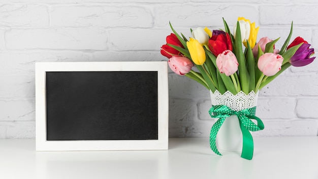 Free photo an empty black frame with tulips vase on desk against white brick wall