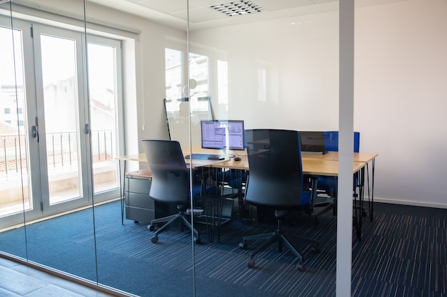 Empty boardroom behind glass wall. Meeting room with conference table, shared desk for team and workplaces. Trading graphs on monitor. Office interior or commercial real estate concept