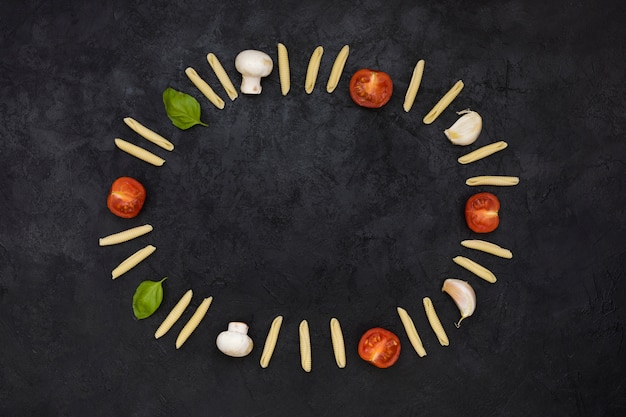 Free photo an empty circular frame formed with uncooked garganelli pasta; tomatoes; mushroom; garlic clove and basil on black textured backdrop