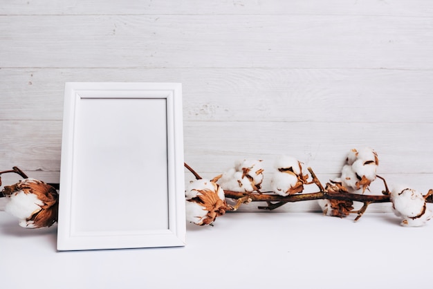 An empty white frame in front of cotton flower stem on desk against wooden background