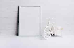 Free photo an empty white frame near the bicycle on white desk against wooden wall