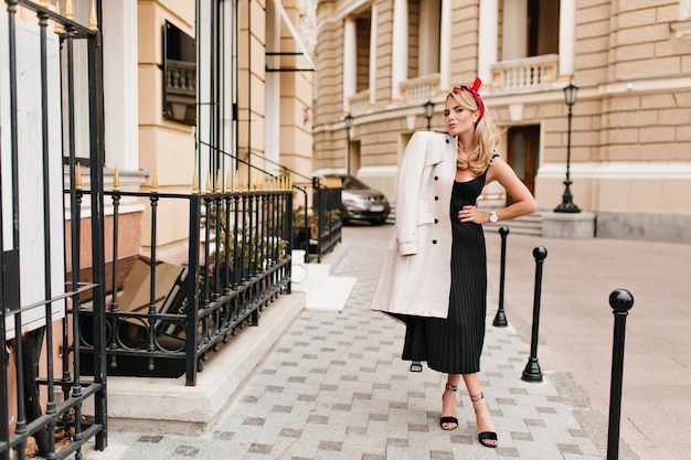 Free photo enchanting fair-haired lady in stylish black dress and high heel shoes posing outdoor in morning. lovely blonde girl in elegant attire spending time on the street and smiling.