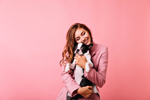 Free Photo enchanting girl with long hairstyle embracing french bulldog with eyes closed. indoor portrait of carefree ginger female model holding cute puppy and laughing.