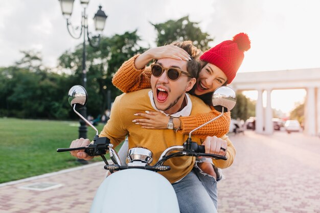 Enchanting lady in red hat touching boyfriend's forehead with smile while he drives scooter