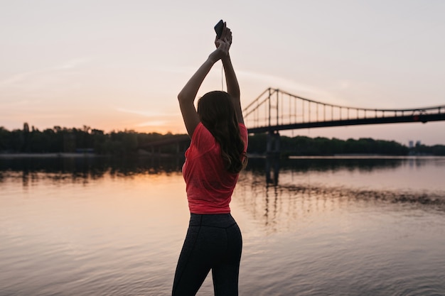 Free photo enchanting shapely girl standing with hands up and looking at river. outdoor photo from back of amazing female model relaxing after training near lake.