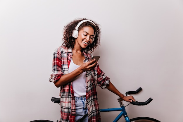 Enchanting stylish woman in headphones reading phone message. Well-dressed african girl with bicycle holding smartphone.