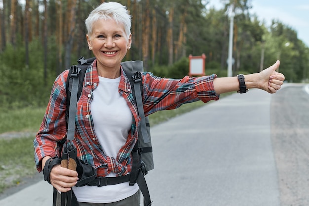 Energetic female pesnioner in active wear standing on road with rucksack behind her back hitchhiking making sign with thumbs up gesture, signaling that she needs a ride.