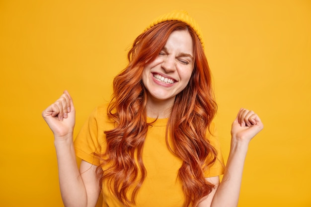 Free Photo energetic upbeat millennial girl smiles happily clenches fists from joy celebrates successful day has natural red wavy hair wears hat casual t shirt 