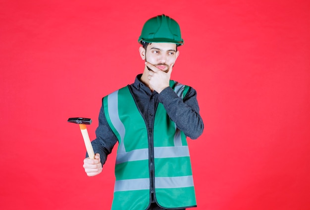 Engineer in green uniform and helmet holding a wooden ax and thinking. 