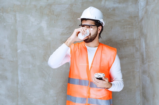 Engineer man in white helmet and protective eyeglasses holding an alarm clock and drinking a cup of tea.