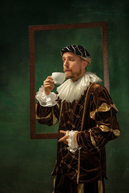 Free Photo enjoy aroma. portrait of medieval young man in vintage clothing with wooden frame on dark background. male model as a duke, prince, royal person. concept of comparison of eras, modern, fashion.