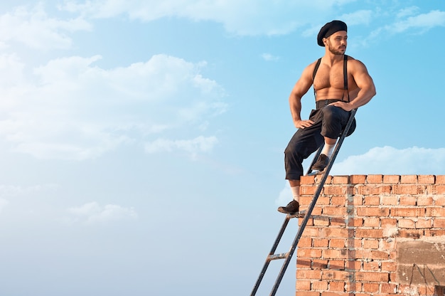 Free photo enjoying outdoors. horizontal shot of a sexy shirtless construction worker sitting on a ladder looking away joyfully blue skies on the background