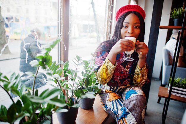 Enthusiastic african american woman in trendy coloured outfit with red beret chilling in cozy cafe with cup of cappucino at hands