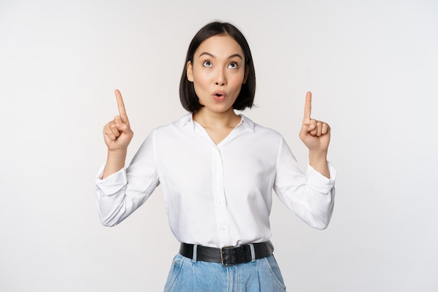 Enthusiastic asian business woman pointing looking up with happy smiling face showing company logo or banner standing over white background