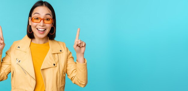 Enthusiastic asian girl in sunglasses points fingers up shows banner or logo on top stands over blue background Copy space