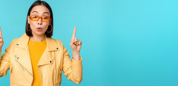 Enthusiastic asian girl in sunglasses points fingers up shows banner or logo on top stands over blue background