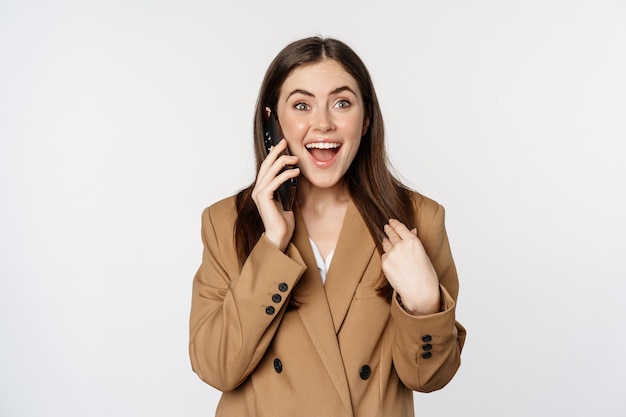 Enthusiastic businesswoman talking on mobile phone, reacting amazed and happy to call, recieve great news, standing over white background