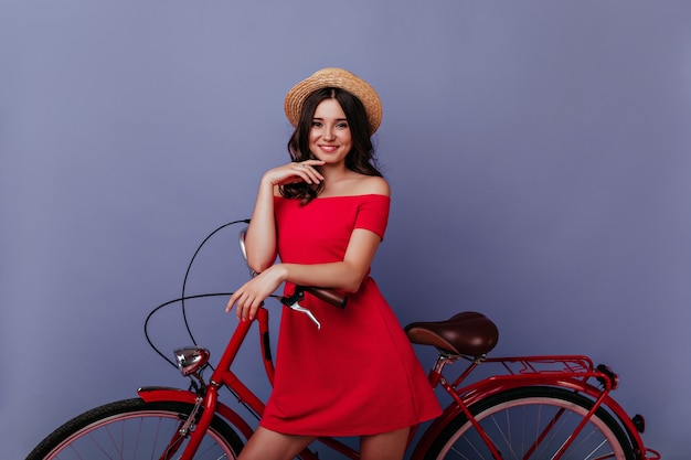 Enthusiastic caucasian girl standing near bike with pleased smile. Indoor photo of gorgeous brown-haired woman enjoying photoshoot on purple wall.