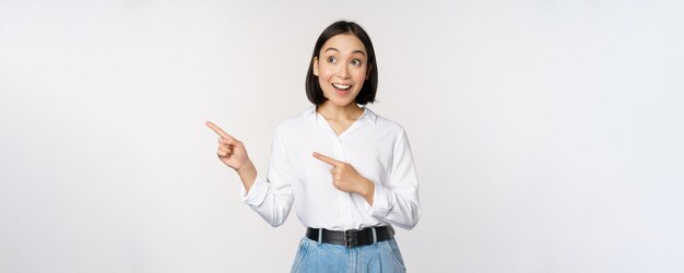 Enthusiastic korean girl pointing fingers left female student pointing and looking left with happy smile showing company logo or banner white background