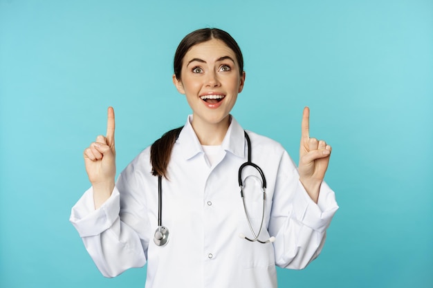 Enthusiastic medical worker, young woman doctor in white coat, stethoscope, showing advertisement, pointing fingers up, standing over torquoise background