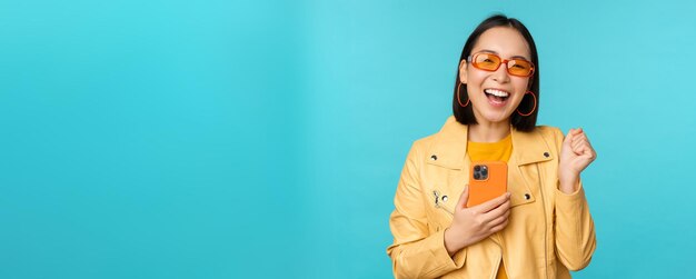 Enthusiastic smiling asian girl in sunglasses holding mobile phone and dancing laughing with smartphone standing over blue background