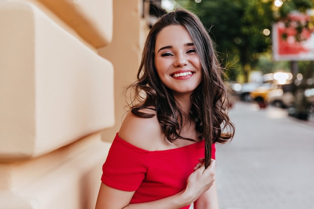 Enthusiastic white woman playing with her wavy hair while standing on the street. Good-humoured female model in red dress posing in city with smile.