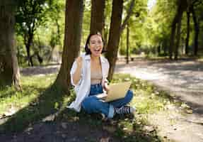 Free photo enthusiastic young asian girl sitting with laptop beside tree in green sunny park celebrating triump