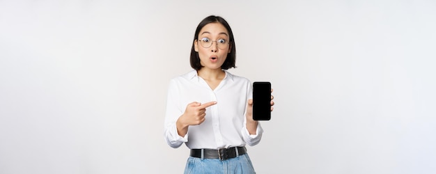 Enthusiastic young asian woman pointing finger at smartphone screen showing advertisement on mobile phone white background