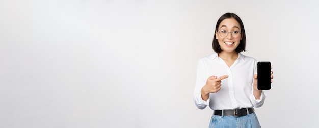 Enthusiastic young asian woman pointing finger at smartphone screen showing advertisement on mobile