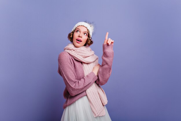 Enthusiastic young lady with short hair posing in long scarf. Indoor photo of fascinating european woman in winter outfit isolated on purple wall.