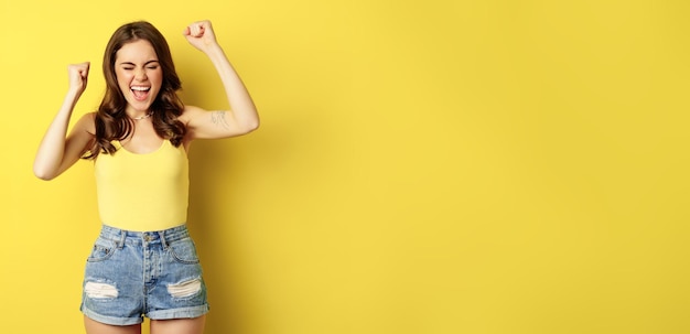 Free photo enthusiastic young woman shouting and cheering rejoicing raising hands up and screaming in joy chanting standing over yellow background