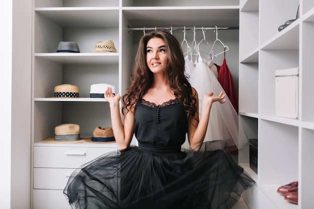 Free photo enthusiastic young woman standing in dressing room, wardrobe and thinking, has contemplative look. her beautiful black dress hovers in the air. she has long curly brown hair.