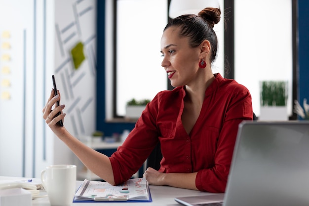 Entrepreneur in casual videocall conference using smartphone. Smiling businesswoman in red shirt using mobile phone in startup business office. Startup manager looking at content on social media.